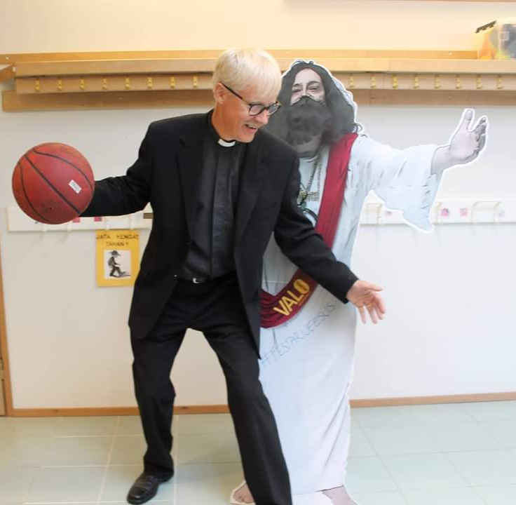 Pahvinen suuri Jeesus-hahmo, pappi päällä ja koripallo kädessä näyttelee pelaavansa pahvi Jeesuksen kanssa